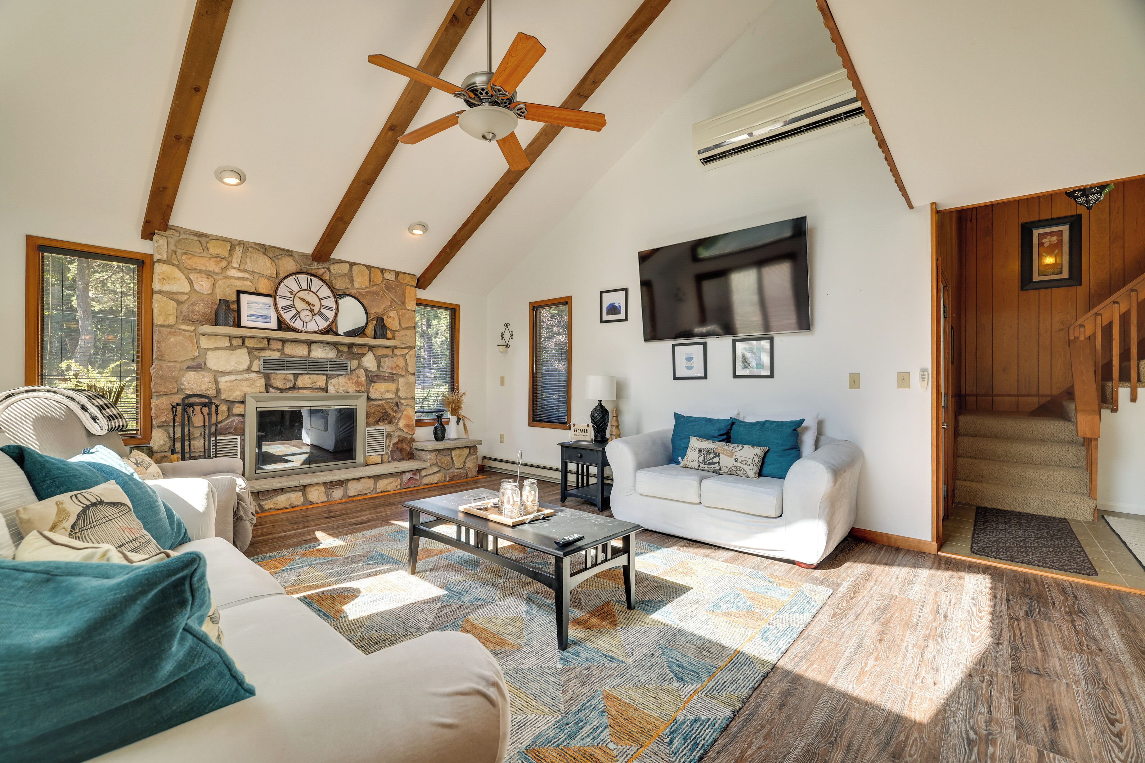 A spacious living room with a stone fireplace, a ceiling fan, a large TV, and a staircase leading to an upper level.