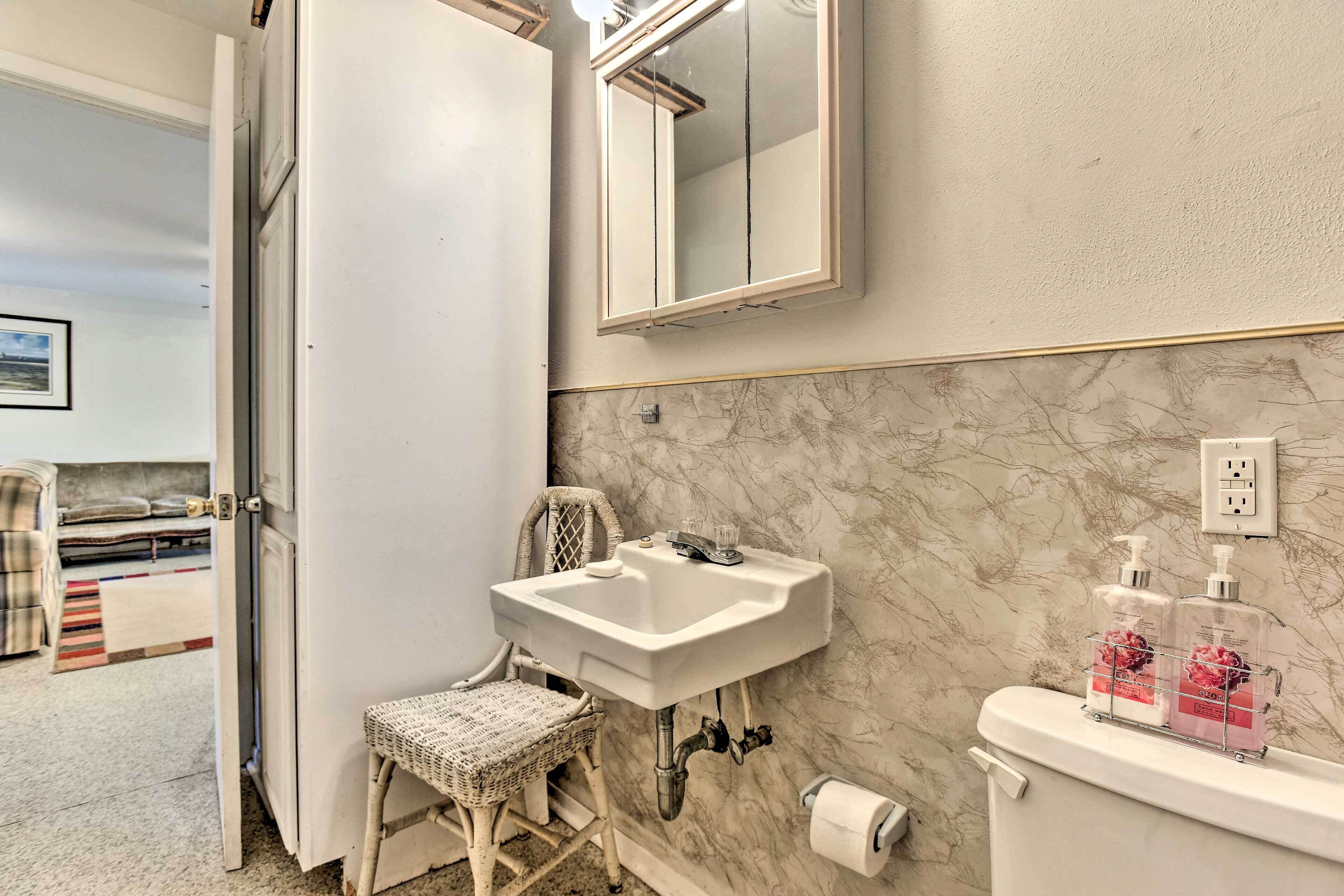 A bathroom with a white sink, a toilet, and a mirror. There is a wicker chair and a small rug on the floor.