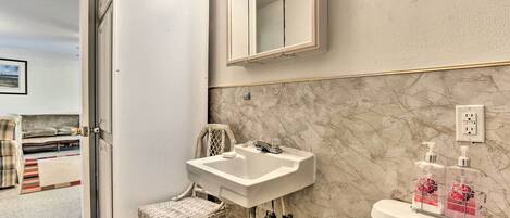 A bathroom with a white sink, a toilet, and a mirror. There is a wicker chair and a small rug on the floor.