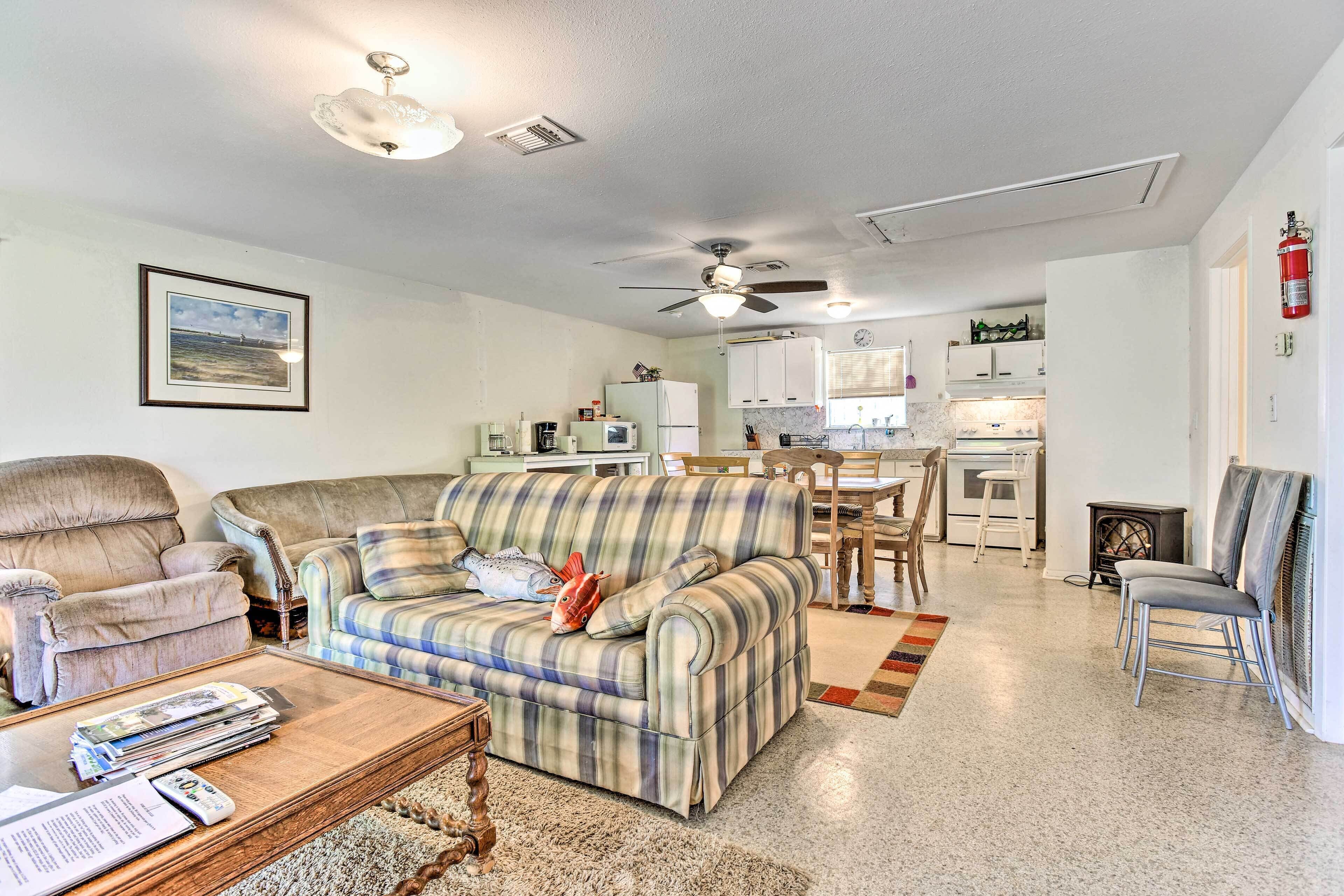 A living room with a sofa, armchair, and coffee table. There is a kitchen in the background with cabinets and appliances.