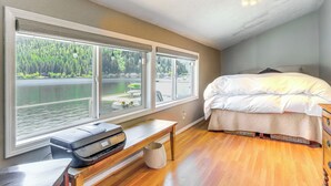 Un dormitorio con una ventana grande que da a un lago, una cama con ropa de cama blanca y un escritorio de madera con una impresora.