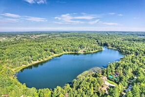 An aerial view of a lake surrounded by dense forest and small buildings, with clear skies above. - Lakefront Hideaway w/ Dock, Kayaks, Fire Pit! (Glennie)