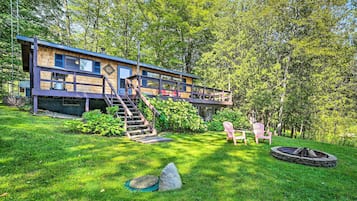 A modern wooden cabin with a deck, a fire pit, and two Adirondack chairs in a lush green yard.