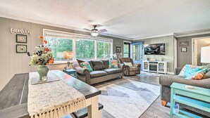 A spacious living room with a large window, a ceiling fan, and a television. - Lakefront Hideaway w/ Dock, Kayaks, Fire Pit! (Glennie)