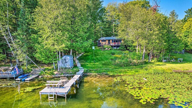 A lakeside cabin with a dock, surrounded by lush greenery and a clear lake with lily pads. - Lakefront Hideaway w/ Dock, Kayaks, Fire Pit! (Glennie)