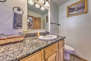 Un bagno con un piano di lavoro in granito, un lavandino bianco, mobili in legno, un porta asciugamani, uno specchio con luci, un quadro e una porta in legno.