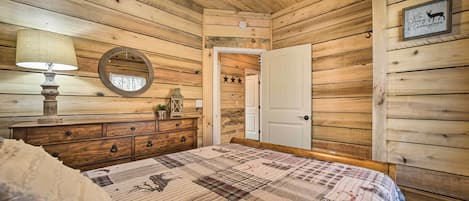 A wooden-paneled bedroom with a bed, a dresser, a mirror, and a door leading to another room.