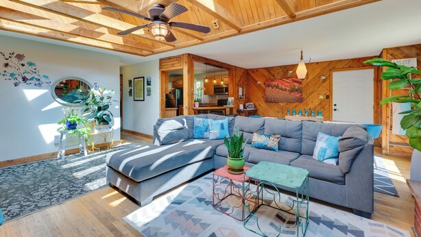 Ein modernes Wohnzimmer mit einem grauen Ecksofa, einem Deckenventilator, Holzbalken und einer durch eine offene Tür einsehbarer Küche.