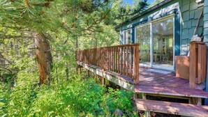 Eine Holzdielen-Terrasse mit Glastür zu einem überdachten Bereich, umgeben von Bäumen und Grünpflanzen.