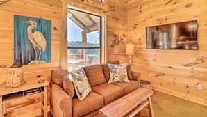 Interior de una cabaña de madera con un sofá, un cuadro de una garza, un televisor y vistas a través de una ventana.