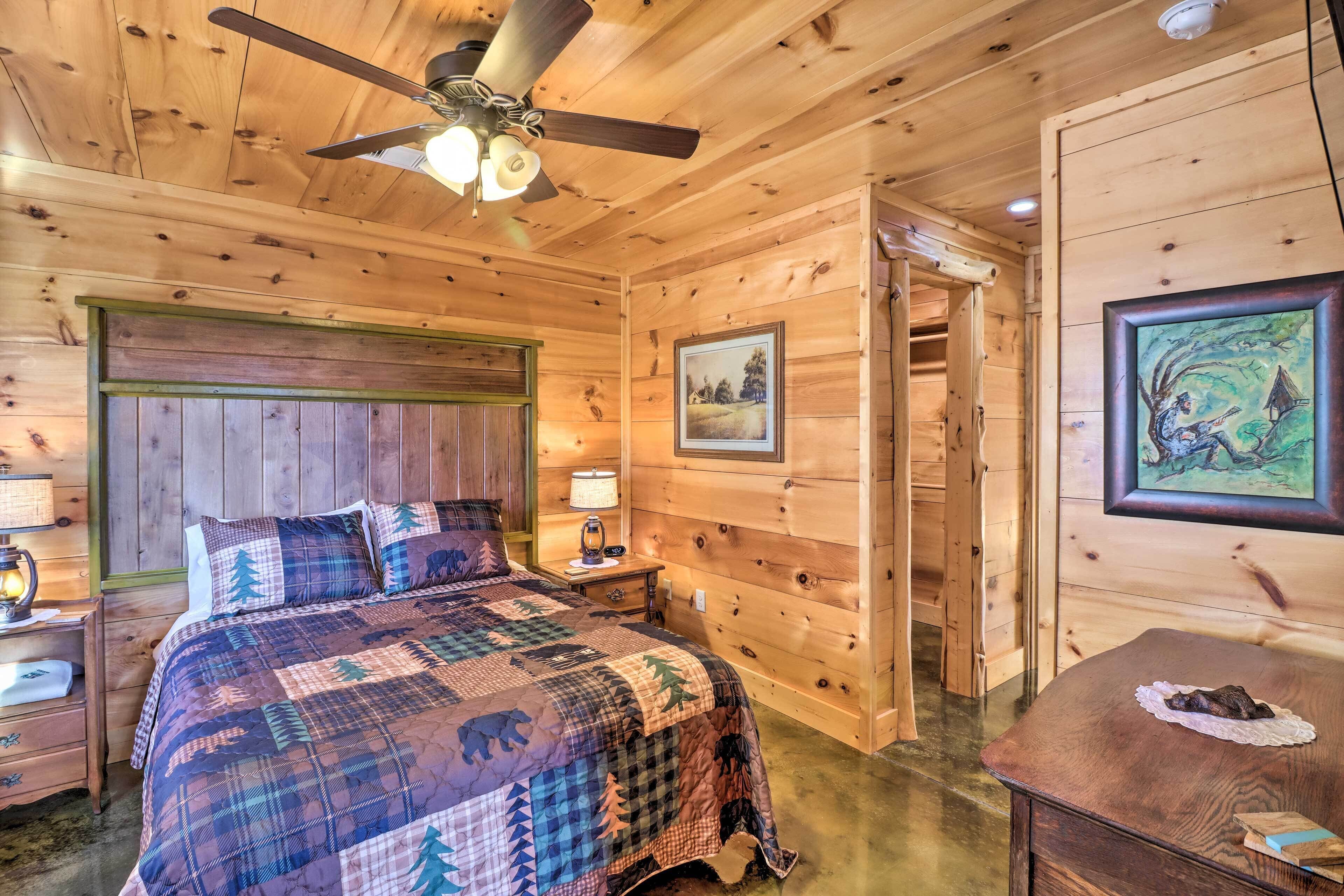 Ein Schlafzimmer einer Holzhütte mit einem Bett, einem Nachttisch, einem Deckenventilator, einem gerahmten Bild und einem kleinen Tisch mit einer Lampe.