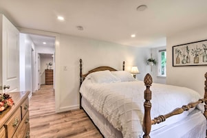 Un dormitorio con estructura de cama de madera, cubrecama blanco, cómoda de madera y un pasillo que conduce a otra habitación.