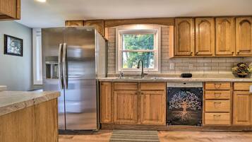 A modern kitchen with wooden cabinets, a stainless steel refrigerator, a dishwasher, and a window with a view of trees.