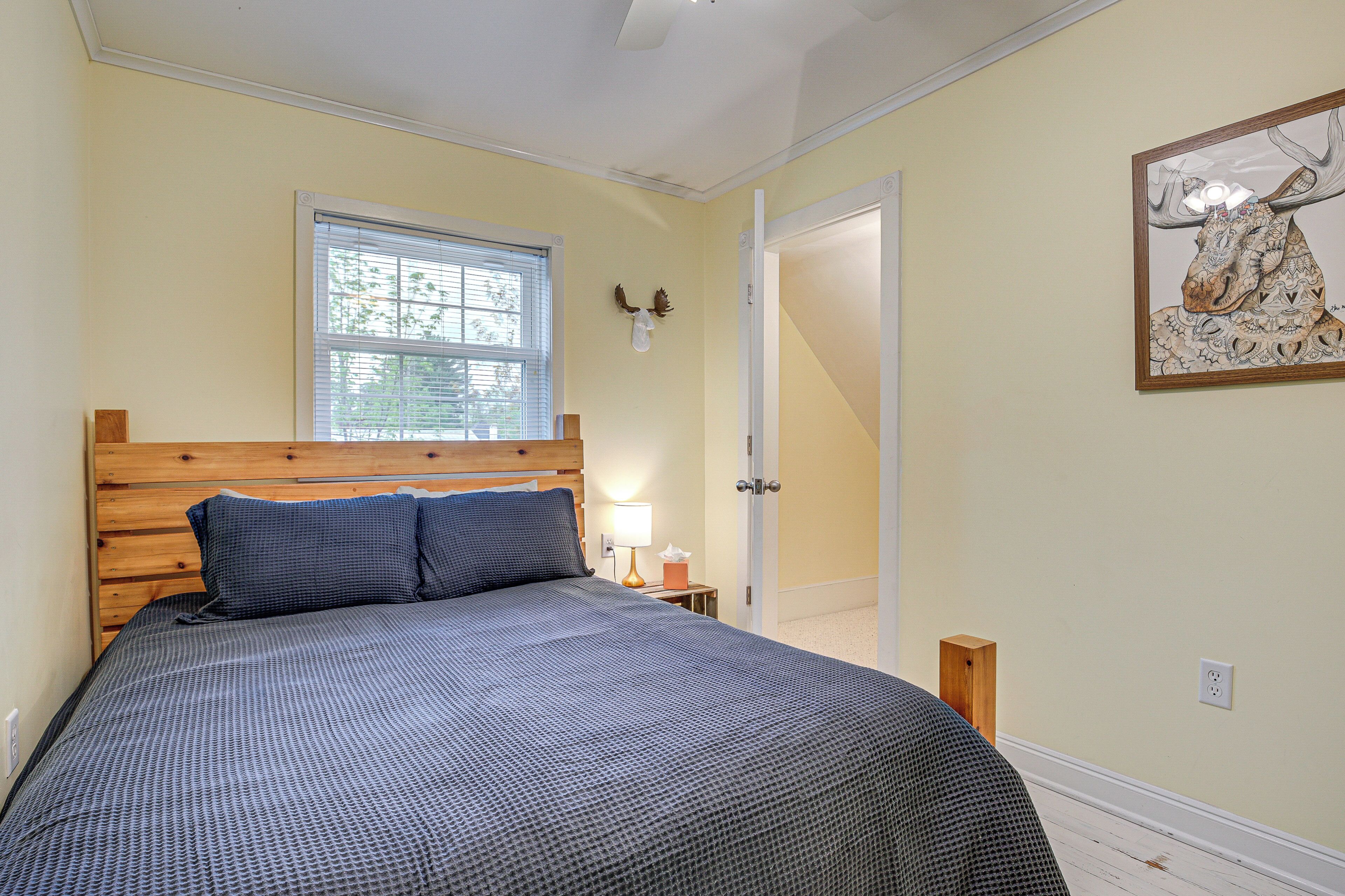 A bedroom with a wooden headboard, a large bed with a patterned bedspread, a window with blinds, a bedside table with a lamp, and a door leading to another room.