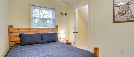 A bedroom with a wooden headboard, a large bed with a patterned bedspread, a window with blinds, a bedside table with a lamp, and a door leading to another room.