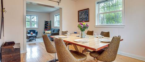 A dining area with a wooden table set for six, surrounded by six woven chairs. There is a vase with flowers on the table, and a framed picture on the wall.