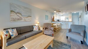 Sala de estar com sofá, poltronas e mesa de centro. Há um quadro na parede, um ventilador de teto e uma cozinha ao fundo.