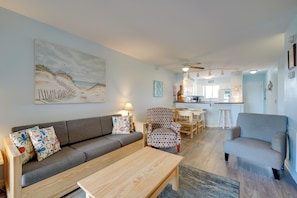 A living room with a sofa, armchairs, and a coffee table. There is a painting on the wall, a ceiling fan, and a kitchen in the background. - Oceanfront Carolina Beach Condo w/ Pool & Views! (Carolina Beach)