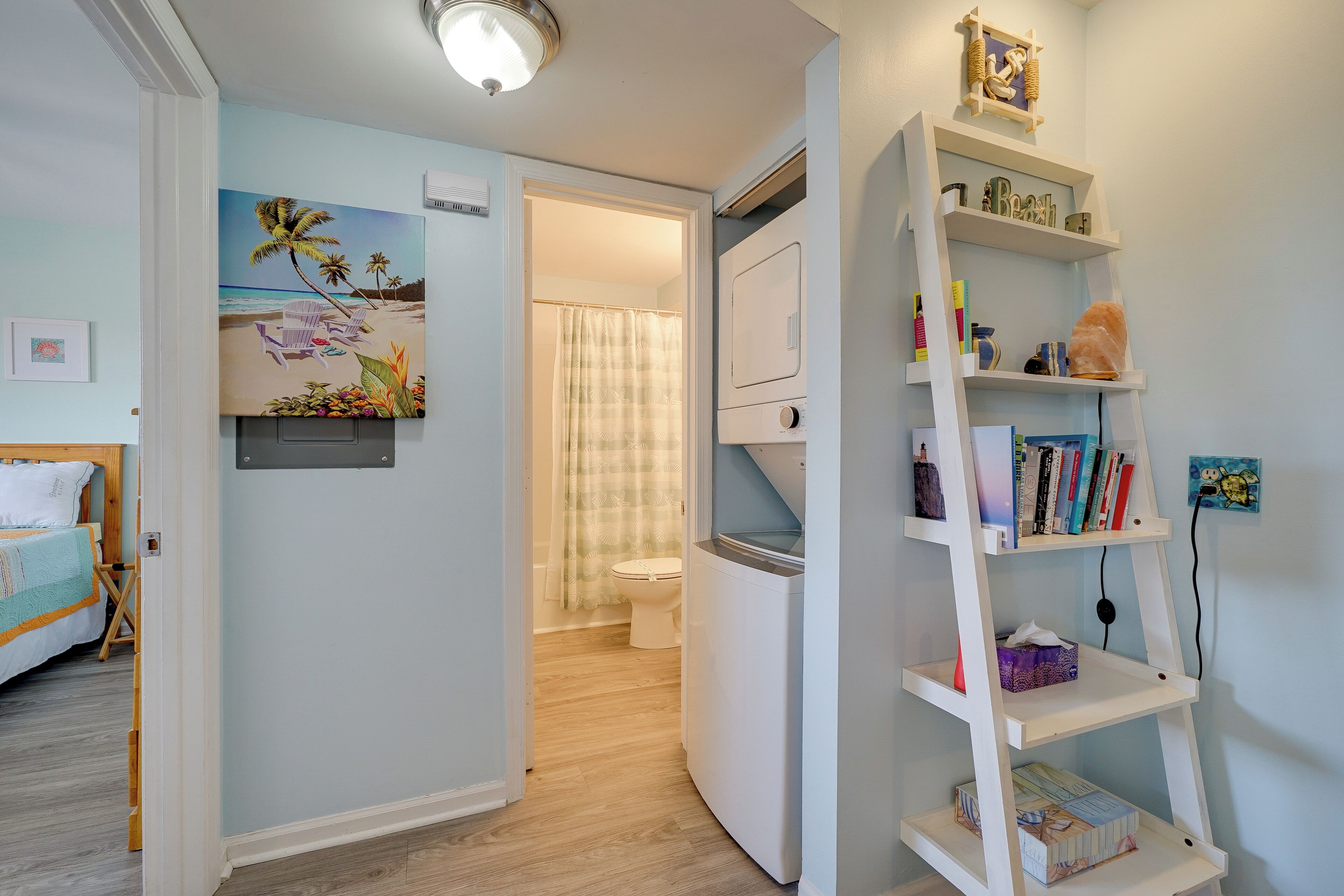 Una habitación con una cama, un cuadro de una escena de playa, un estante blanco con libros y una lavadora blanca.