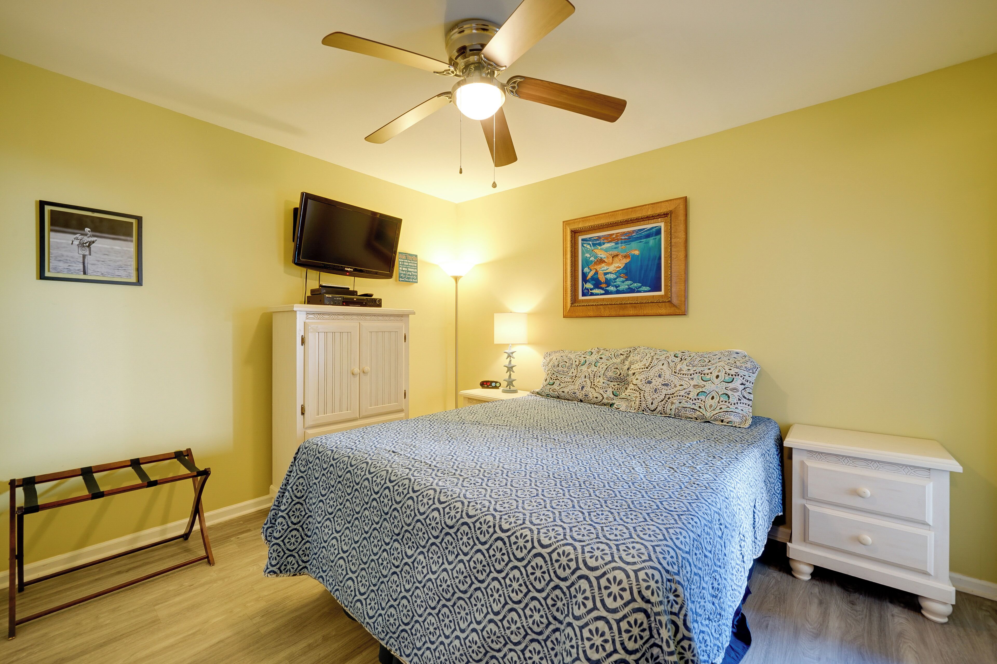A bedroom with a bed, a ceiling fan, a TV, a lamp, a cabinet, and a framed picture on the wall.