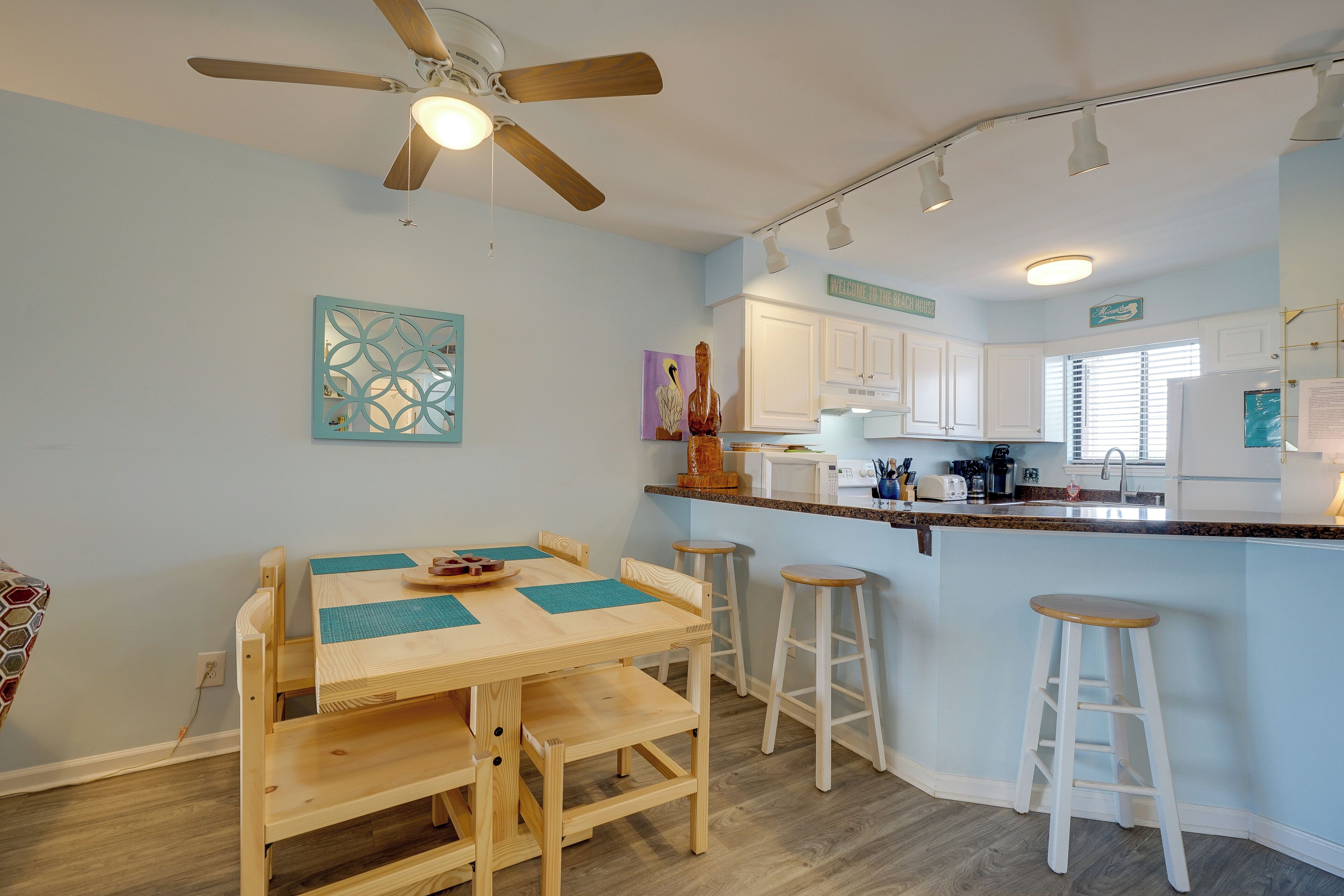 Una cocina moderna con armarios de madera, una isla central con taburetes, un ventilador de techo y un letrero que dice "Welcome to the Beach House".