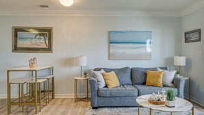 A living room with a sofa, coffee table, and side tables, featuring artwork and a beach scene. - Ocean Views + Resort Pool! N Topsail Beach Condo (Sneads Ferry)
