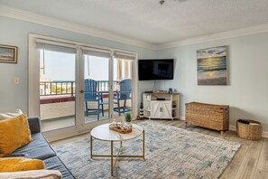 Een woonkamer met een bank, een rond bijzettafeltje, een televisie, een rieten tafeltje en uitzicht door een schuifpui naar buiten.