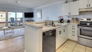 A modern kitchen with a dishwasher, stove, and a large window overlooking a balcony. - Ocean Views + Resort Pool! N Topsail Beach Condo (Sneads Ferry)