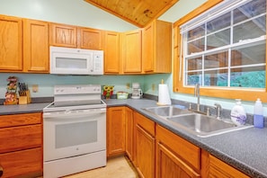 A kitchen with wooden cabinets, a microwave, a stove, a sink, and a window with a view of greenery. - Peaceful Bryson City Cabin - Fire Pit & Hot Tub! (Bryson City)