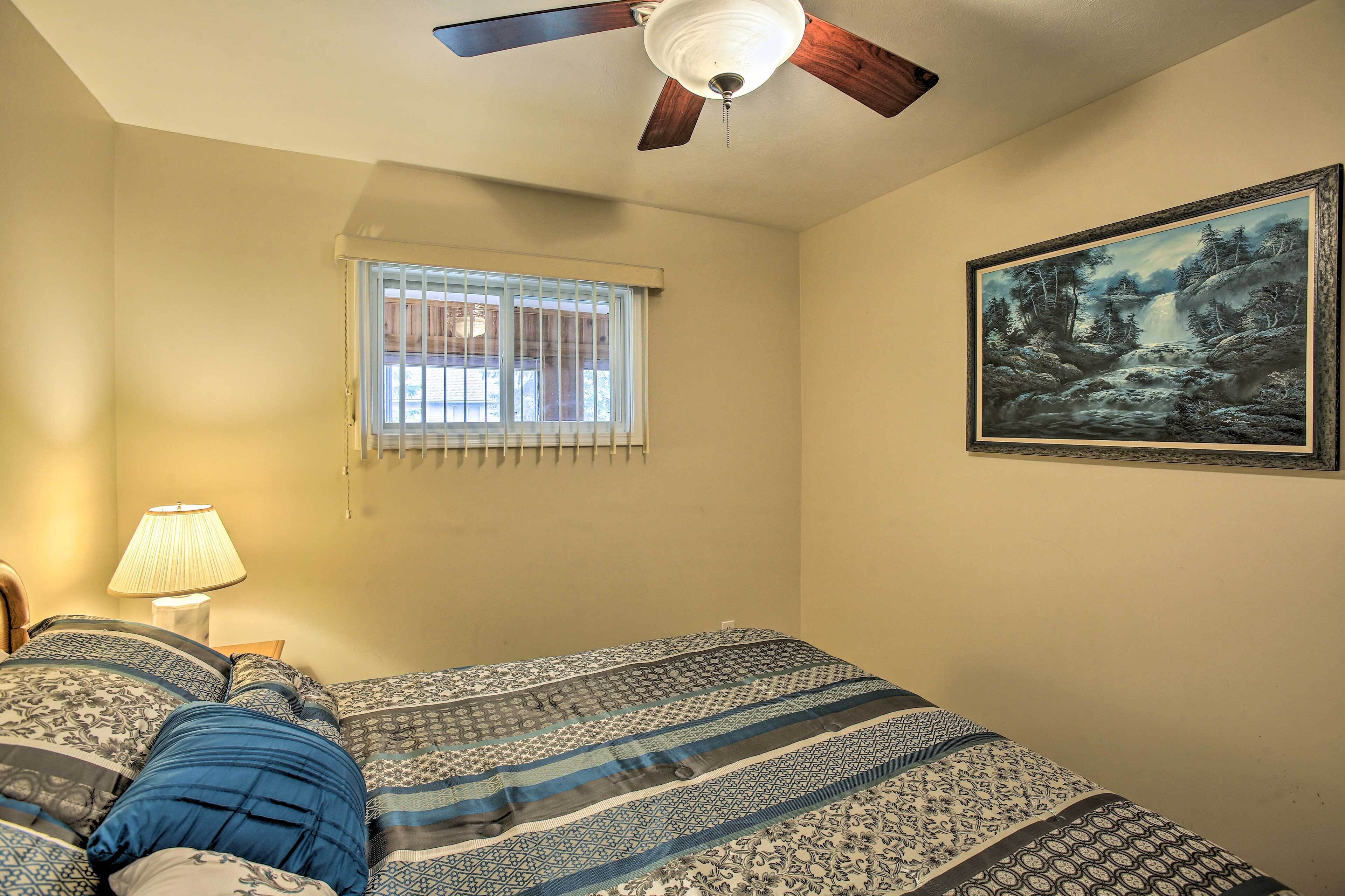 Een slaapkamer met een bed, een plafondventilator, een raam met jaloezieën, een lamp en een lijst met een schilderij aan de muur.
