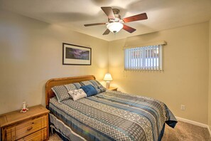 A bedroom with a bed, wooden nightstand, dresser, ceiling fan, window with blinds, and a framed picture on the wall. - Peaceful Lakefront Houghton Lake Property w/ Patio (Prudenville)