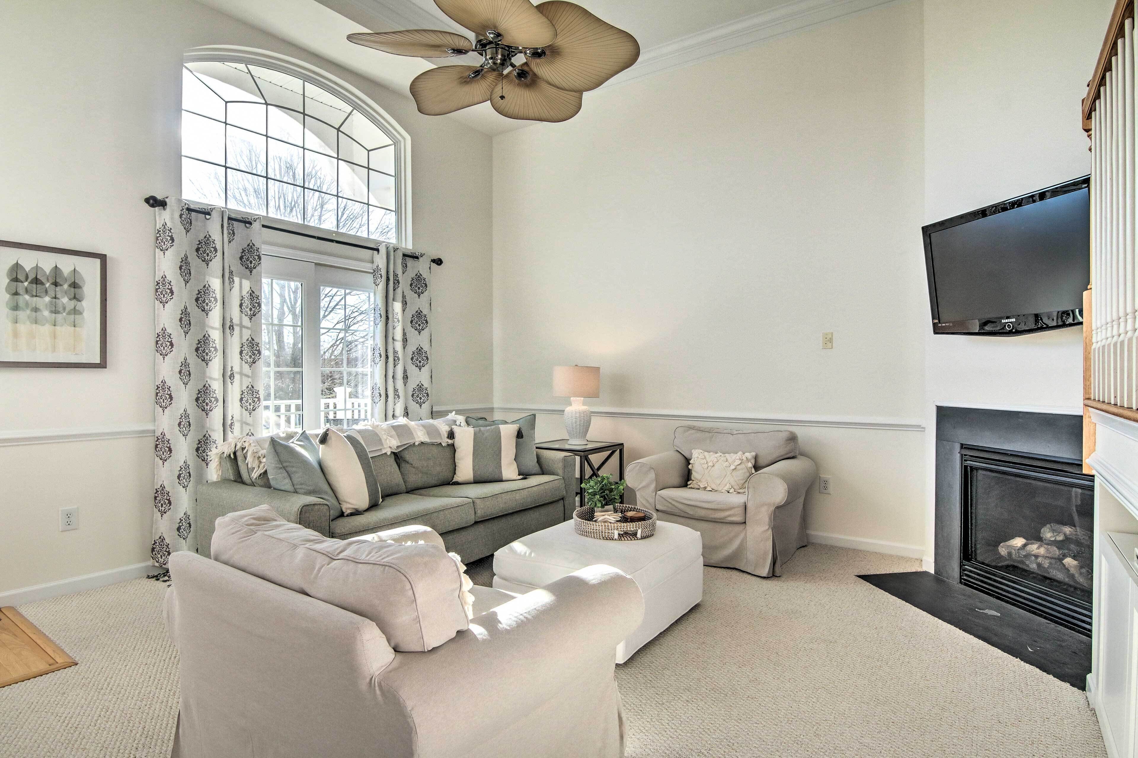 A living room with a fireplace, a large window with curtains, a ceiling fan, and a flat-screen TV mounted on the wall.