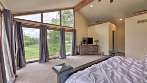 Un dormitorio con una cama grande, televisión, cómoda y vistas a un paisaje verde a través de amplias ventanas.