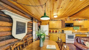 Interior de una cabaña de madera con una mesa de comedor, sillas, un florero con flores, un sofá y una cocina con armarios y electrodomésticos de madera.