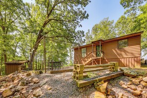 Un chalet rustique avec un escalier en bois menant à une terrasse, entouré d’arbres et bordé d’un chemin de gravier.