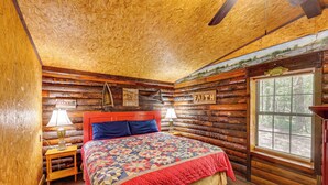 Un dormitorio de cabaña de madera con techo de madera, una cama con colcha roja y azul, una mesita de noche con lámpara, y una ventana con vistas a los árboles.