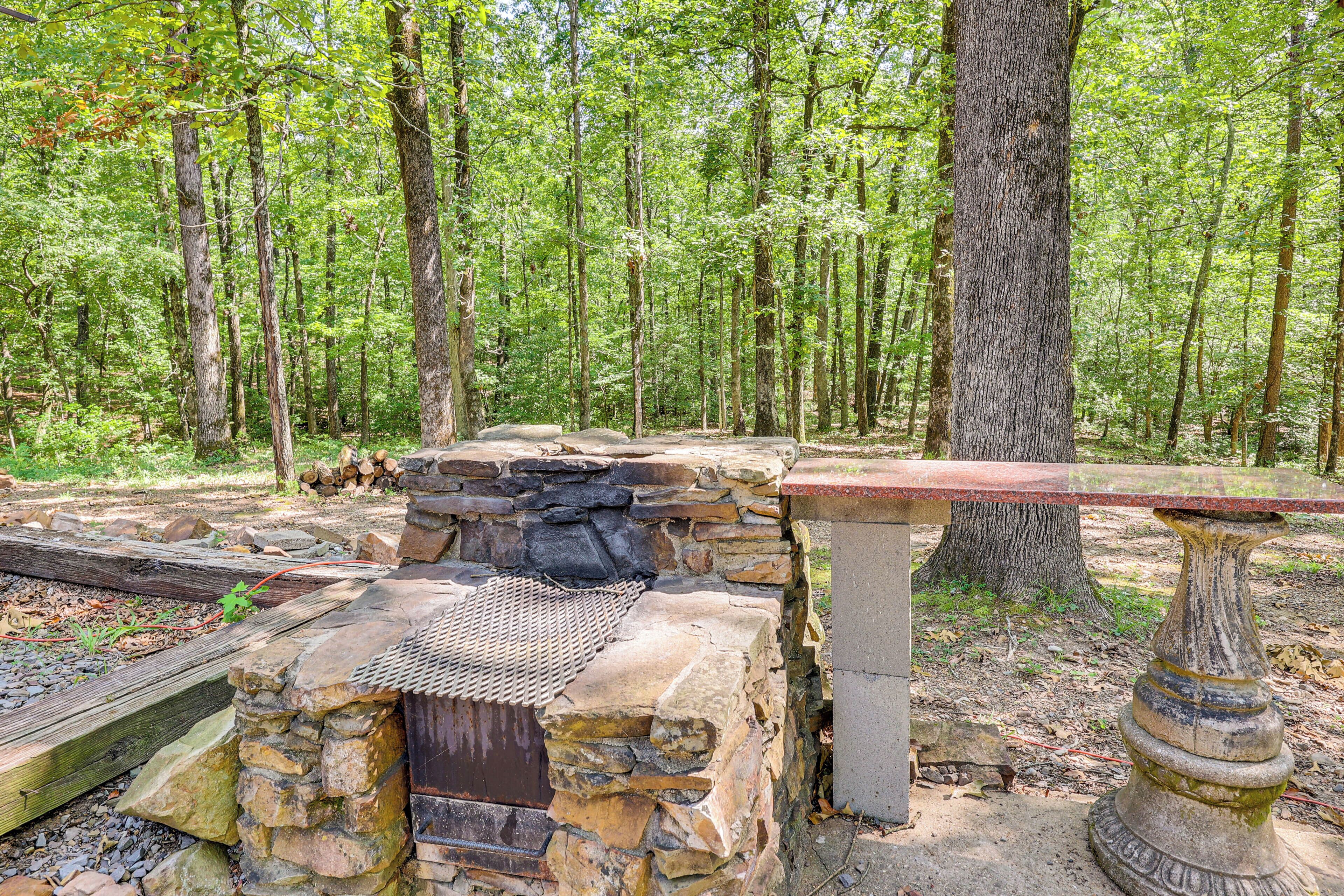 A stone bench and a stone table in a forested area with a small stone fireplace.