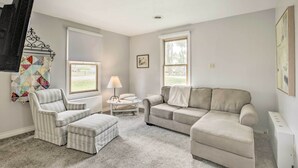 Una sala de estar con un sillón a rayas, un sofá color beige y un reposapiés a juego. Hay una mesita con una lámpara, un colcha en la pared y una ventana con vistas al exterior.