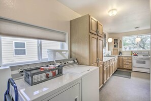 A laundry room with a front-loading washer and dryer, wooden cabinets, a window with blinds, and a kitchen area with a stove and cabinets.