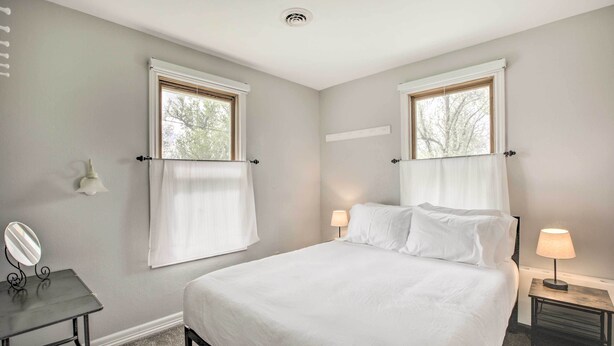 Un dormitorio con una cama grande, dos mesitas de noche con lámparas, un escritorio pequeño con espejo y dos ventanas con cortinas.