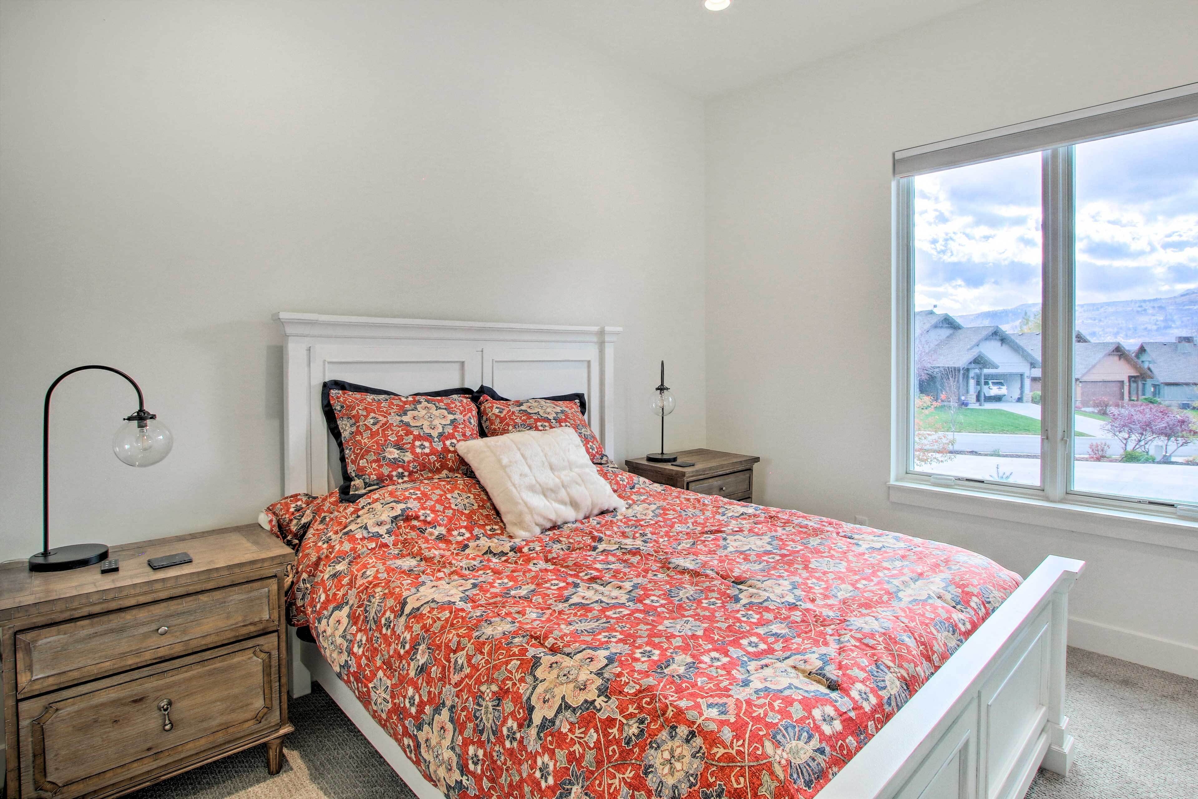 A bedroom with a white bed, floral bedding, a nightstand with a lamp, and a window with a view of houses.