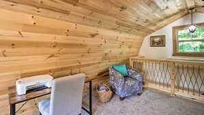 Un cuarto acogedor con paredes y techo de madera, una impresora sobre una mesa, una silla, un banco acolchado y una ventana con vistas a la vegetación.