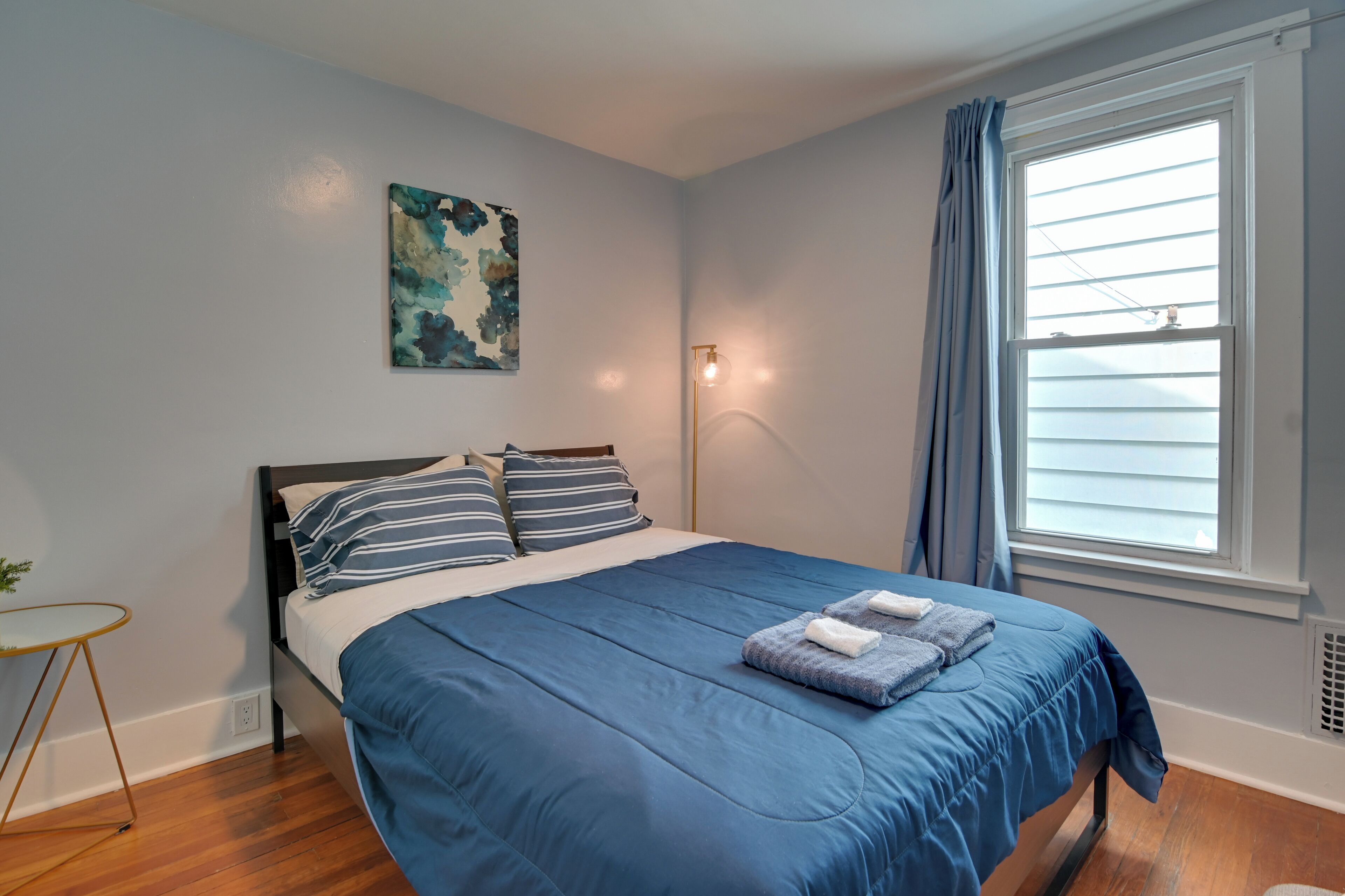 A bedroom with a bed, a bedside table, a lamp, a window with curtains, and a wall art piece.