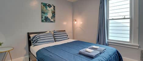 A bedroom with a bed, a bedside table, a lamp, a window with curtains, and a wall art piece.