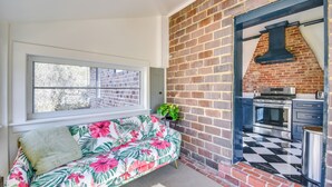 A cozy room with a sofa, a window, a brick wall, and a kitchen area with a checkered floor. - Radiant Gloucester House w/ Private Porch! (Gloucester)