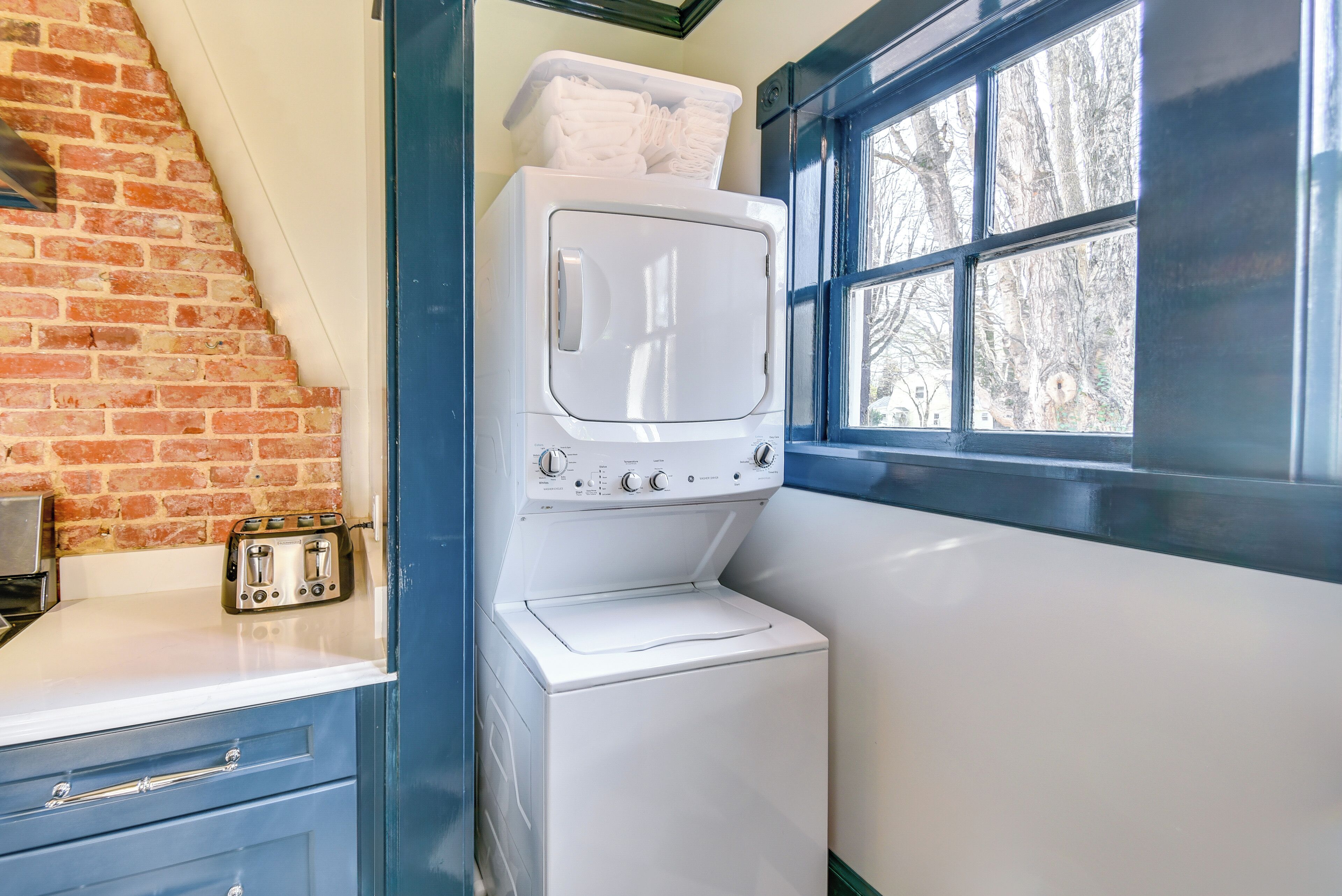 A laundry area with a washing machine and dryer, a brick wall, a window with a view of trees, and a countertop with a toaster.