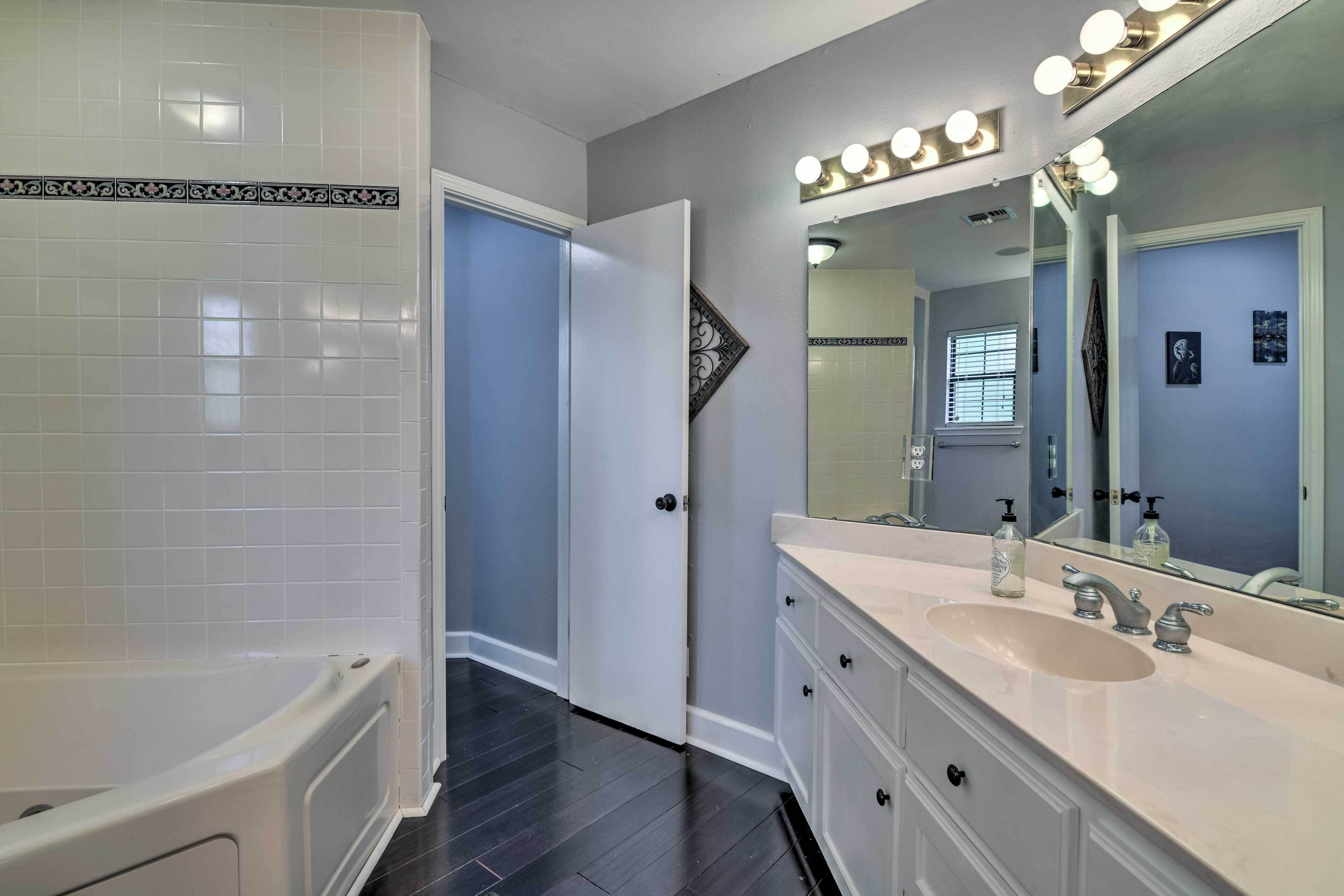 Un baño con bañera, tocador con lavabo y espejo, y una puerta que da a otra habitación.