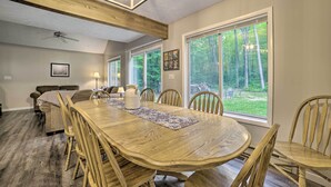 A dining area with a large wooden table, chairs, and a view of a forest through large windows. - Higgins Lake Getaway - Boating, Fish, Golf, Atv! (Roscommon)