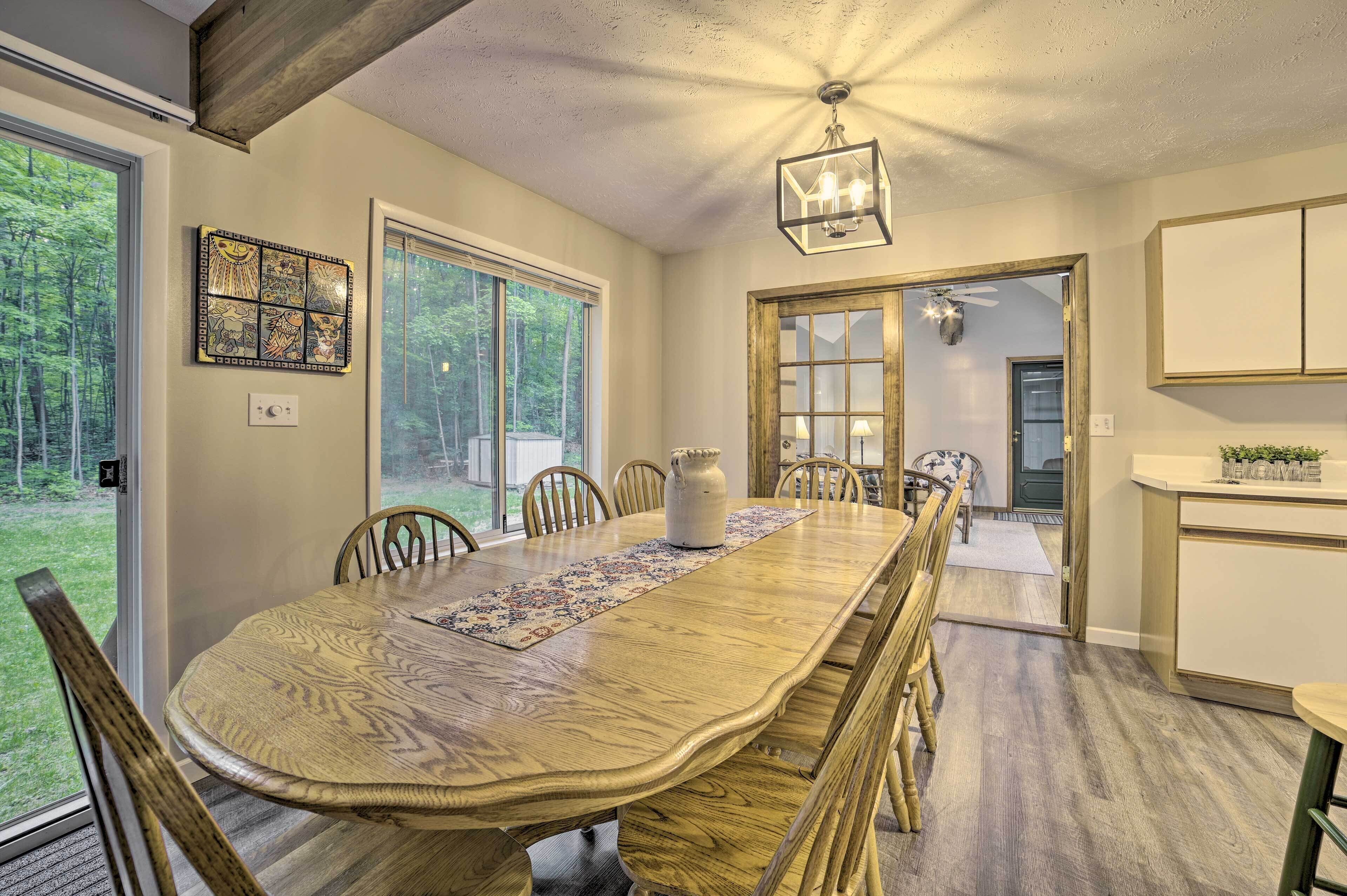 A dining room with a large wooden table, chairs, and a chandelier. There is a door leading to another room, and a window with a view of trees.