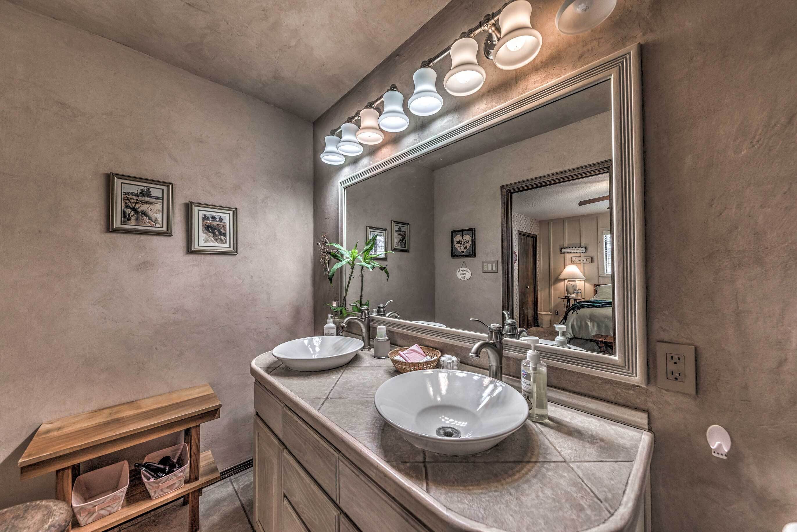 A bathroom with two sinks, a large mirror, and wall-mounted lights. There are framed pictures on the wall and a small shelf with items.